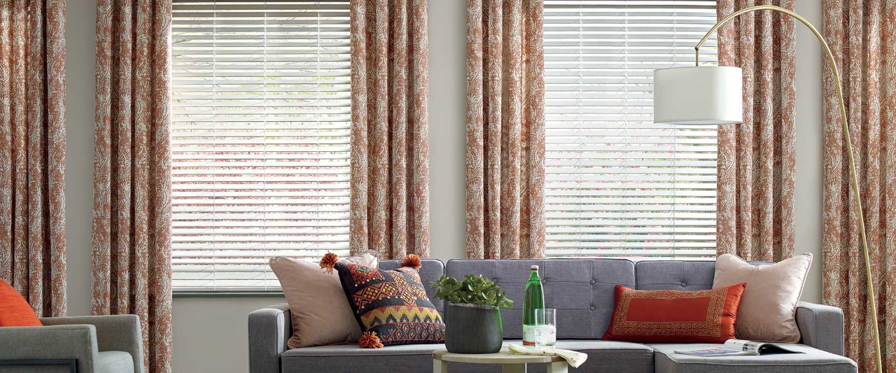Grey couch with orange pillows in front of closed faux wood blinds